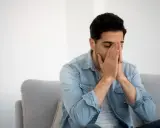 Mann sitzt auf Sofa, Hände vors Gesicht gepresst, Augen geschlossen. Fühlt sich nach dem Rauchen schwindelig.