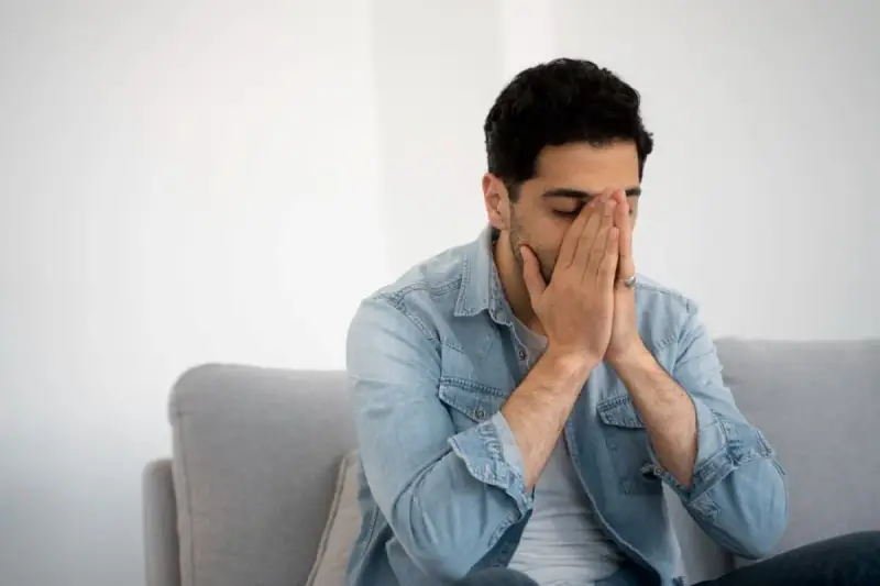Mann sitzt auf Sofa, Hände vors Gesicht gepresst, Augen geschlossen. Fühlt sich nach dem Rauchen schwindelig.